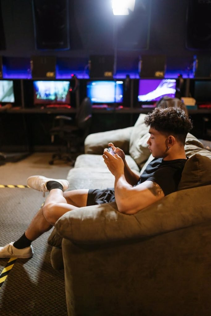 A young man in casual attire relaxes on a sofa playing games on his smartphone at an internet cafe.